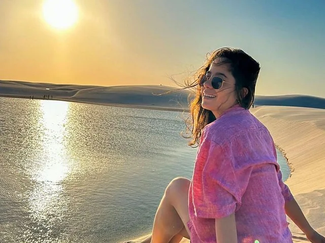 Frau in rosa Hemd und Sonnenbrille, auf Sanddünen am Wasser sitzend