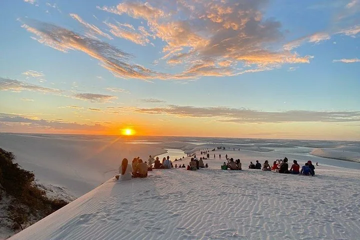 Eine Gruppe von Menschen sitzt auf Sanddünen und beobachtet den Sonnenuntergang über Lagunen.